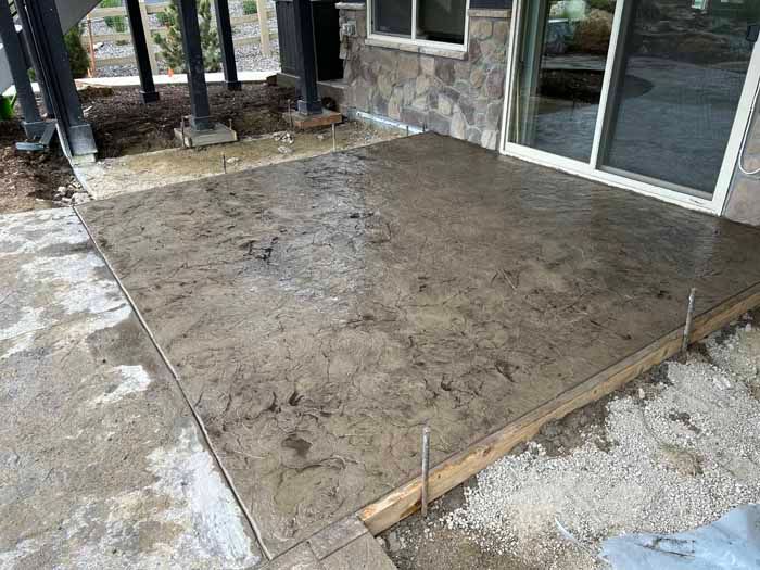 Wet, stamped concrete patio with wooden frame next to a house with stone siding and sliding glass door.