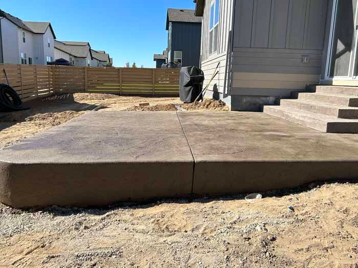 A freshly poured concrete patio next to a house with steps. A wooden fence is in the background.