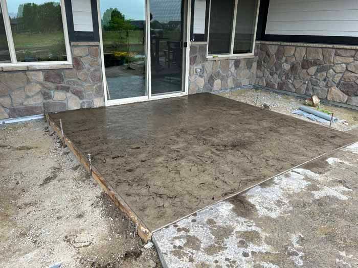 Newly poured concrete patio, brown with mottled texture, next to a house with stone siding.