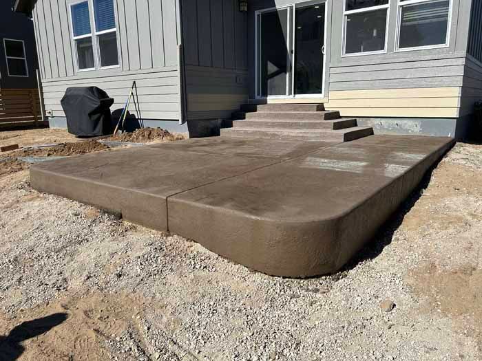 Newly poured brown concrete patio with steps leading to a house, gravel surrounding.