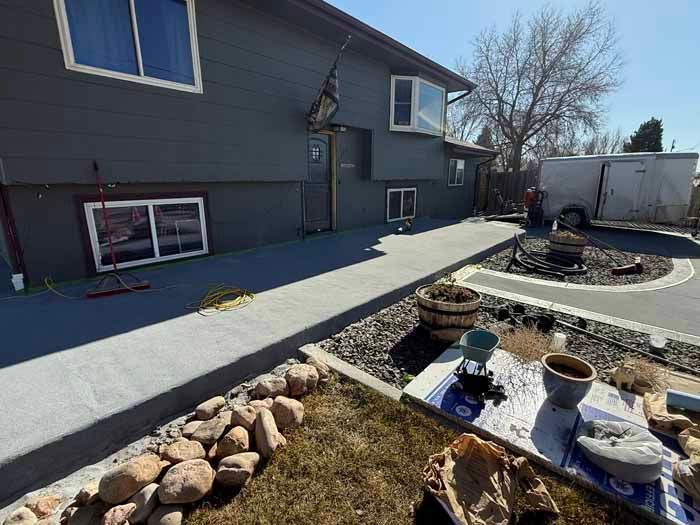 Gray house with concrete walkway, rocks, and a trailer on a sunny day.