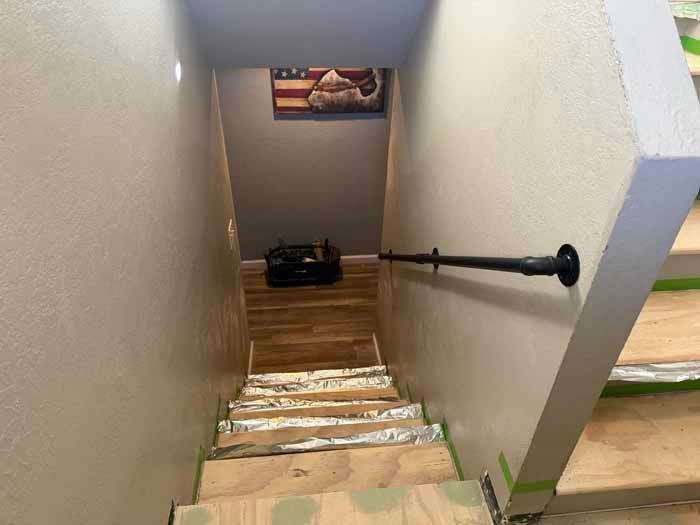 Stairwell with wooden steps, a black handrail, and beige walls, leading downward to a dark room with an American flag.