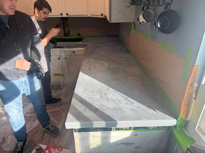 Two men working on a concrete countertop in a kitchen; light grey, cabinets, mugs hanging, walls taped.