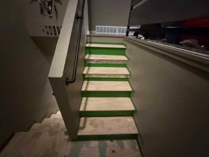 Staircase with green-painted edges and plywood steps, handrail on the left, leading downwards into a basement.