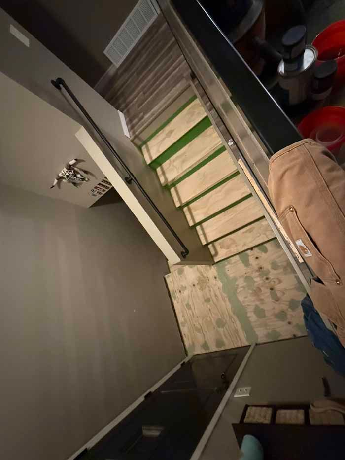 View from above a staircase with visible unfinished wooden steps. A person leans against a rail near some paint cans.