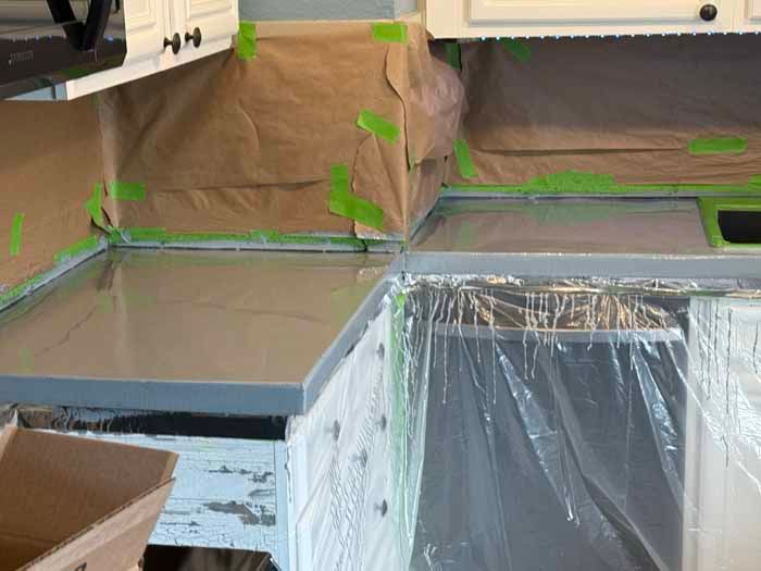 Kitchen countertop being painted, covered in masking paper and plastic, with green tape.