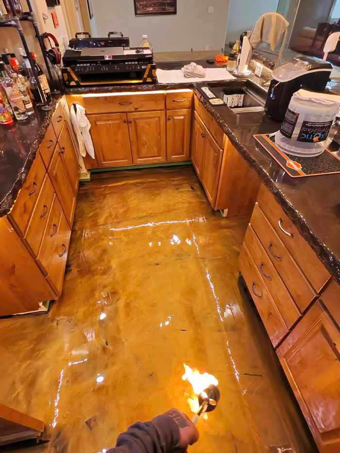 Person using a torch on a shiny, gold epoxy floor in a kitchen with wooden cabinets and dark countertops.