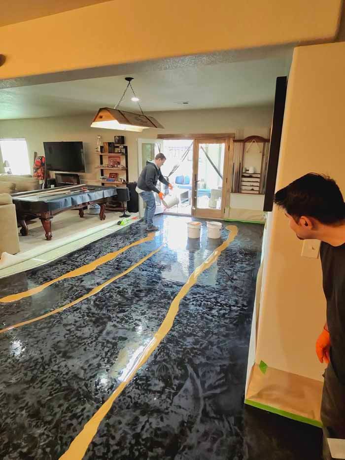 Man applying epoxy floor coating in a room. Black floor with gold accents, another man watches.