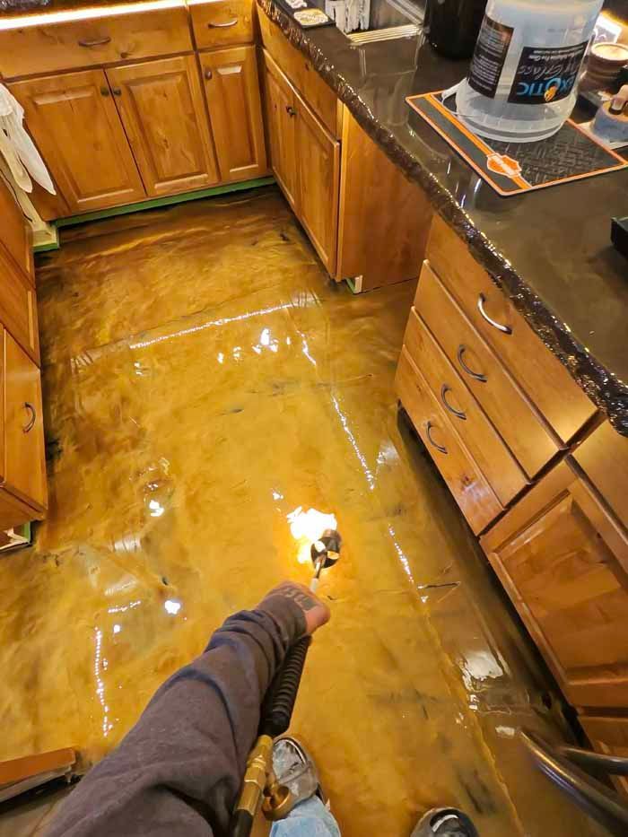 Person torching metallic gold epoxy floor in a kitchen with wooden cabinets.