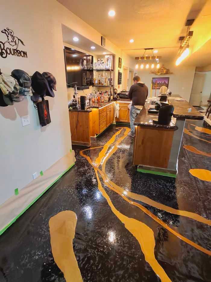 A person near a wet, epoxy-finished bar. Black floor with gold streaks, wood cabinets, and bar stools.
