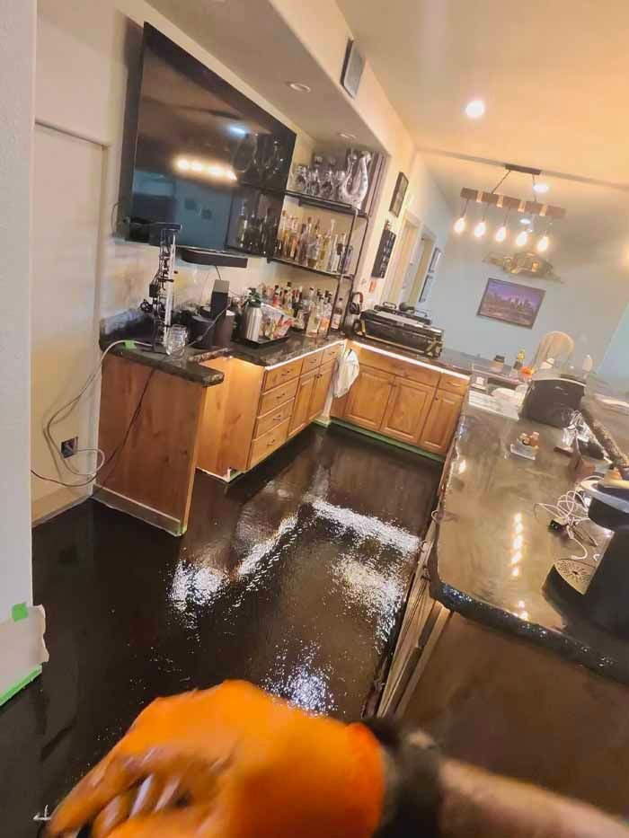 Kitchen with black floor, wood cabinets, and bar. Orange-gloved hand in foreground.