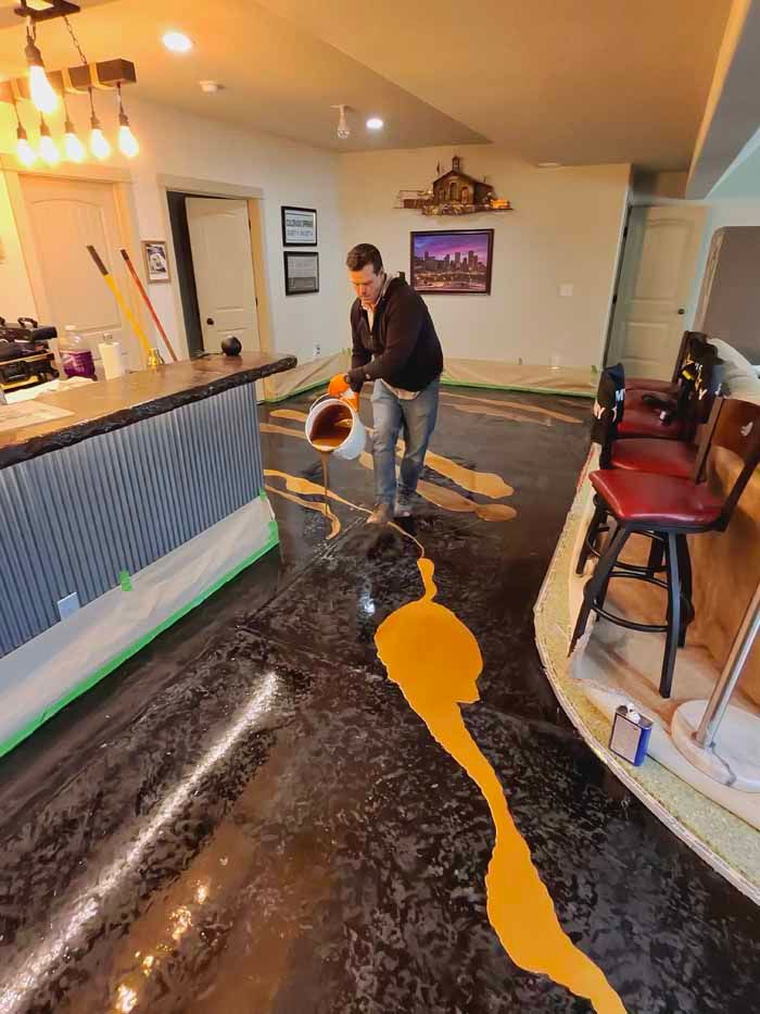 Man pouring orange epoxy onto a black epoxy floor in a basement.