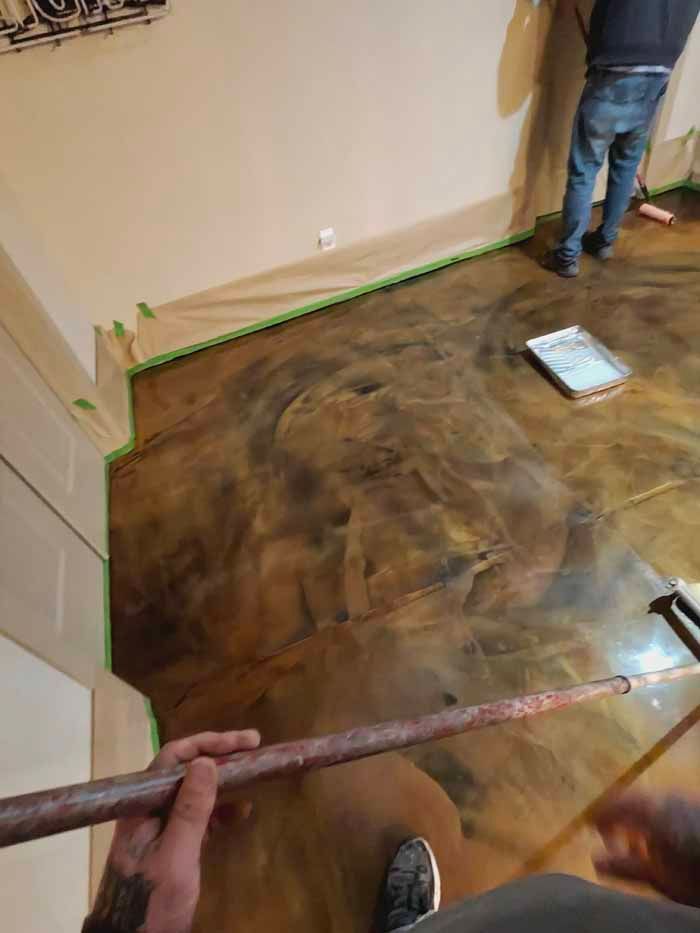 Person applying epoxy to a brown and gold floor. Walls are taped, person holds a tool.