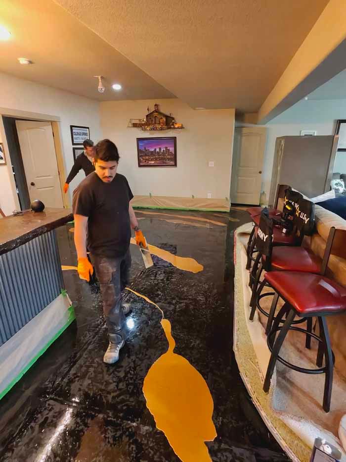 Person applying yellow epoxy to a black floor in a home. The floor is near a bar with red stools. Another person in the background.