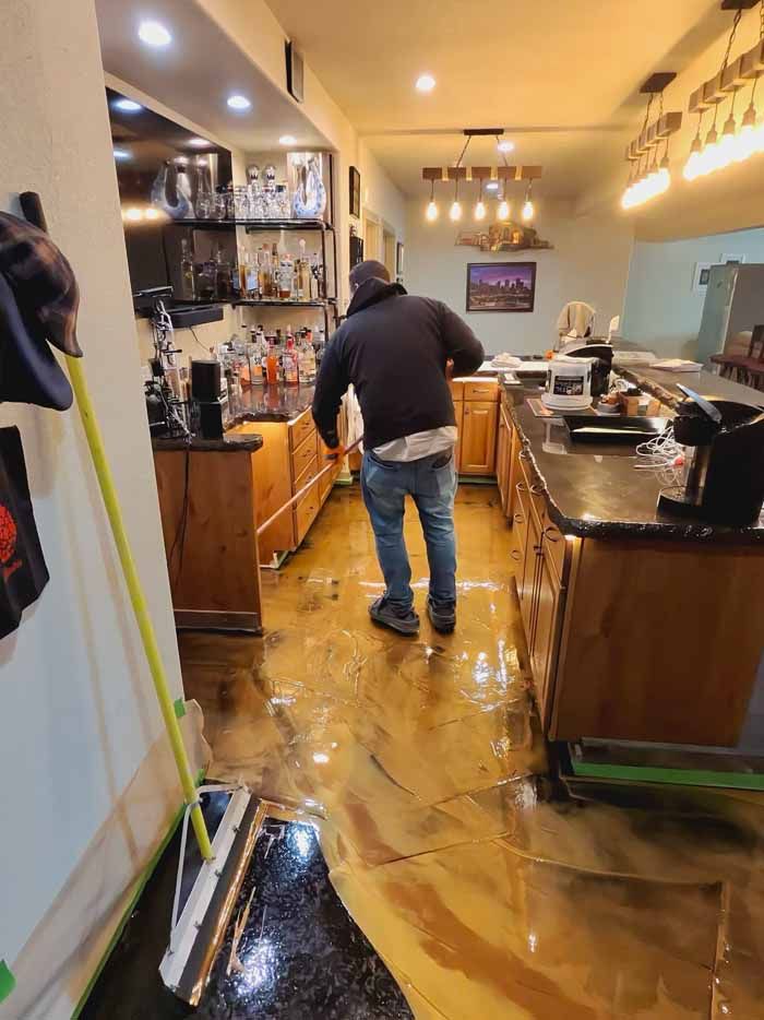 A person mops a wet, metallic-gold epoxy floor in a bar. Wooden cabinets and liquor bottles are visible.