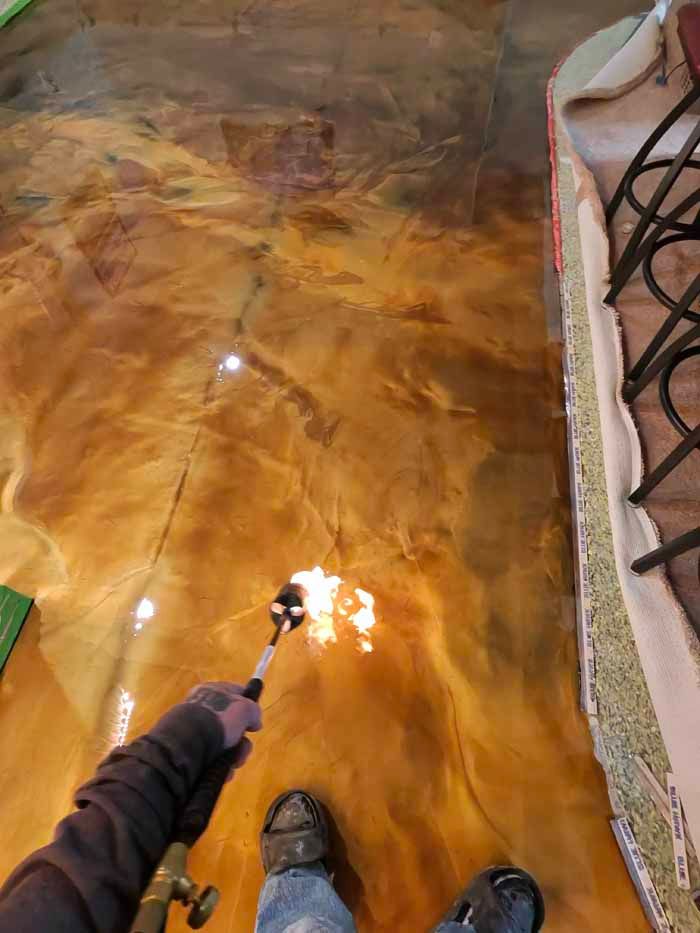 Person using a torch on a gold-colored epoxy floor; close-up shot of feet and floor.