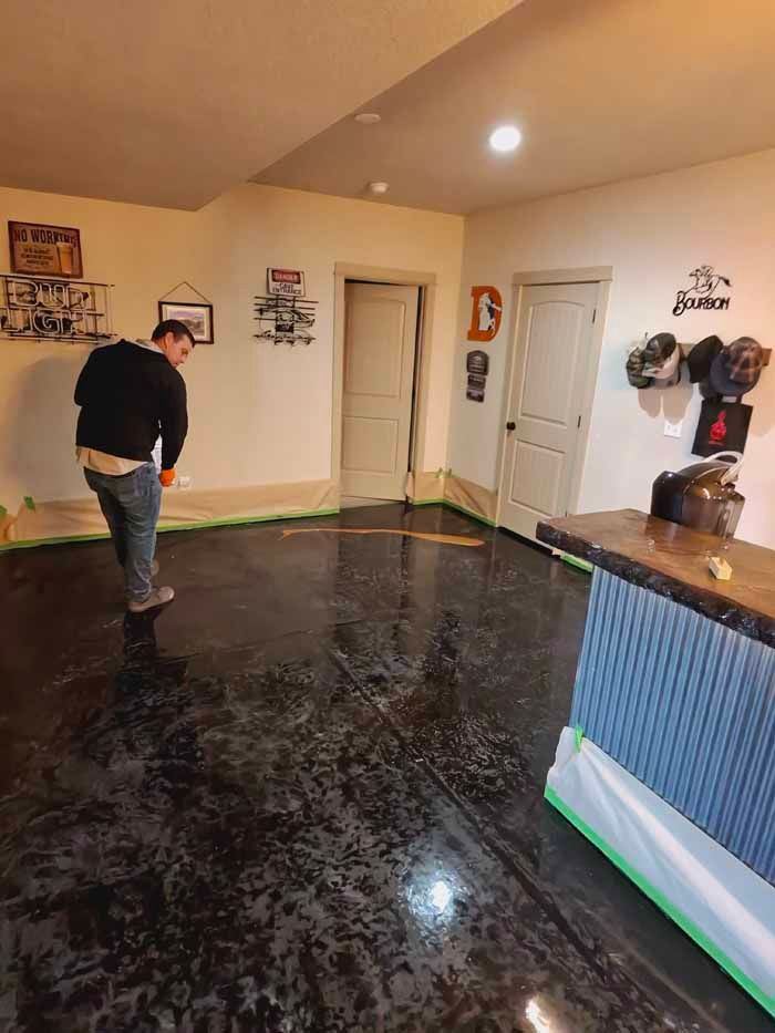 Person applying sealant to a dark, shiny floor in a room with a bar and doors.