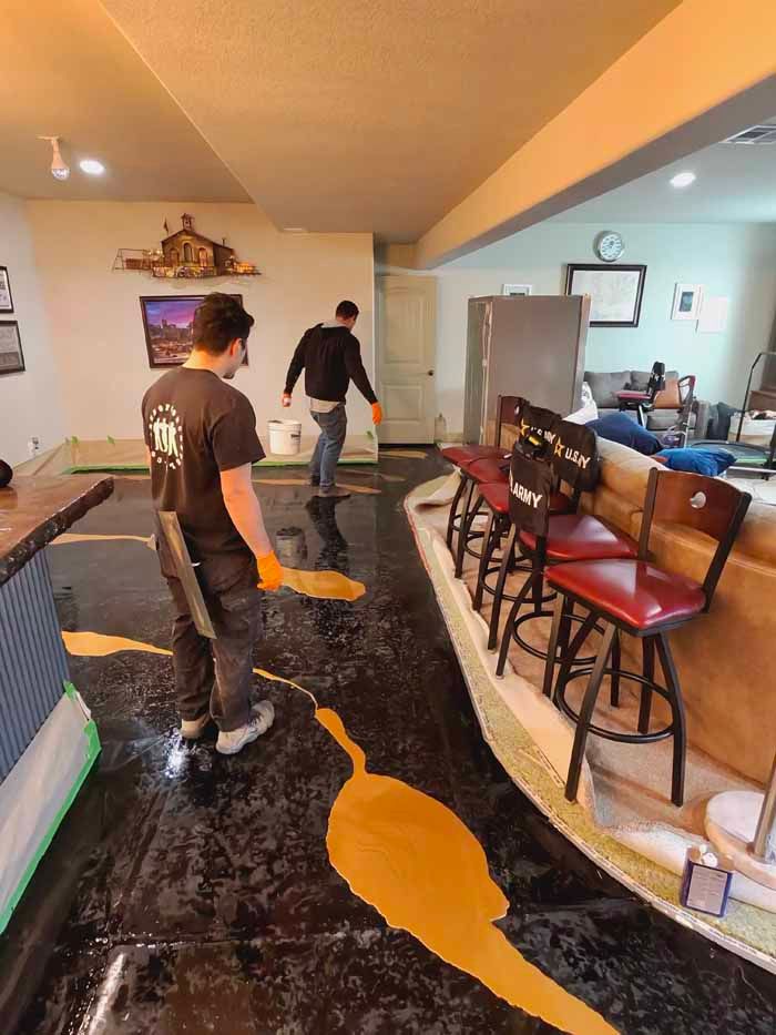 Two people applying yellow epoxy to a black floor in a room with a bar.