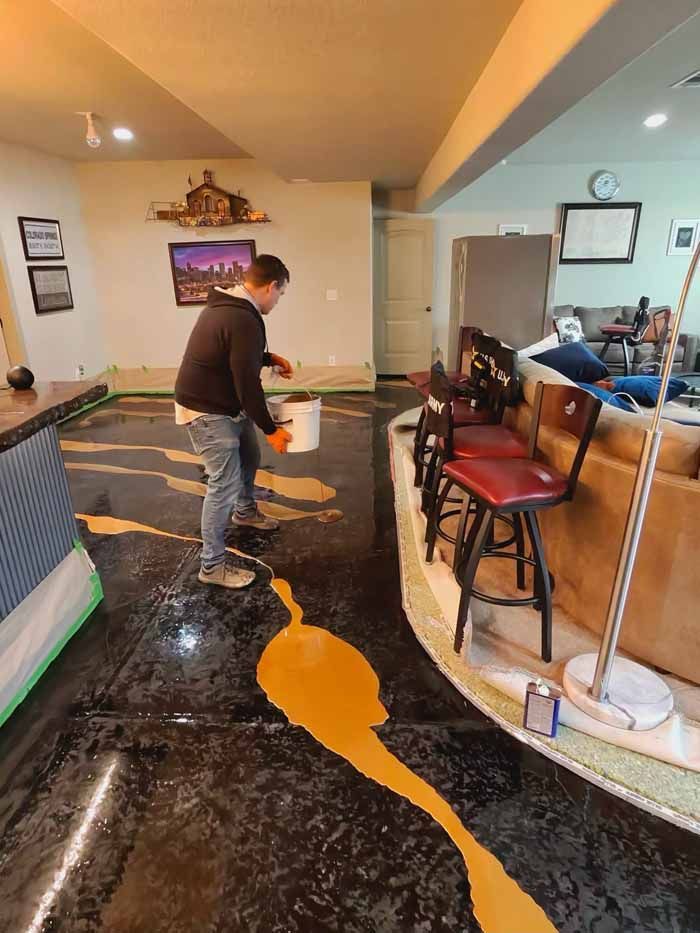Man pouring orange material onto a dark floor, preparing to coat it. The space is a basement with a bar.