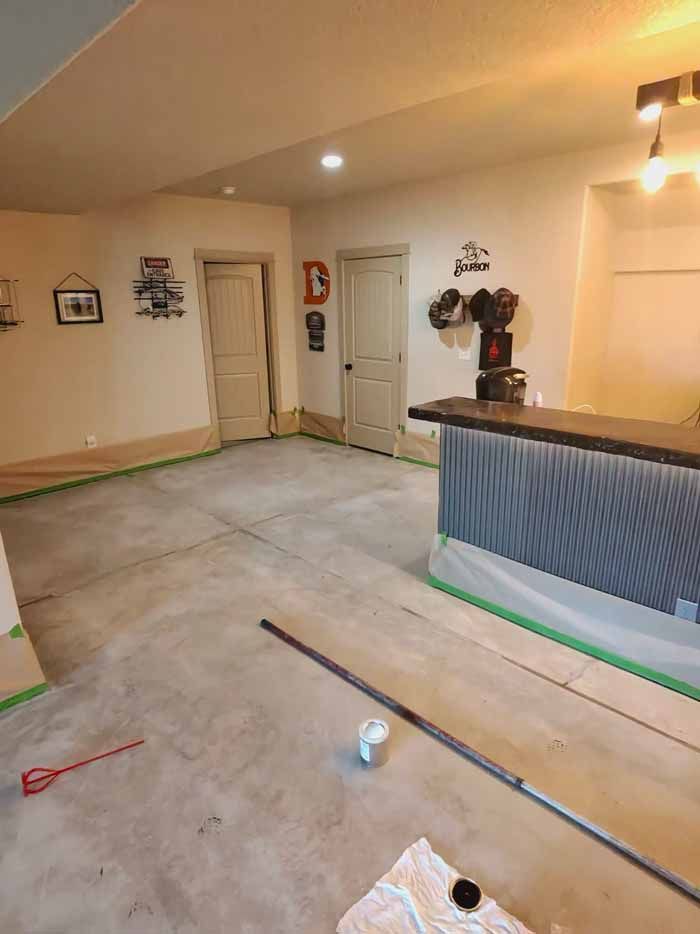 Basement interior with concrete floor, bar, doors, and painting supplies. Green tape protects walls.
