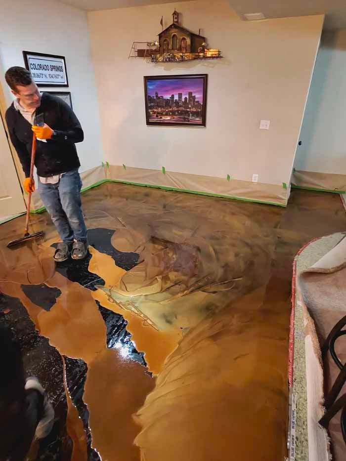 A man applies epoxy to a floor. The floor has a metallic, multi-colored finish. Room has artwork and is taped off.