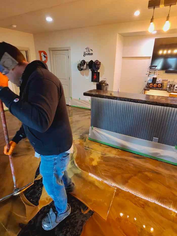 Man applying epoxy flooring with a squeegee inside a room with a bar. The floor is gold and brown.