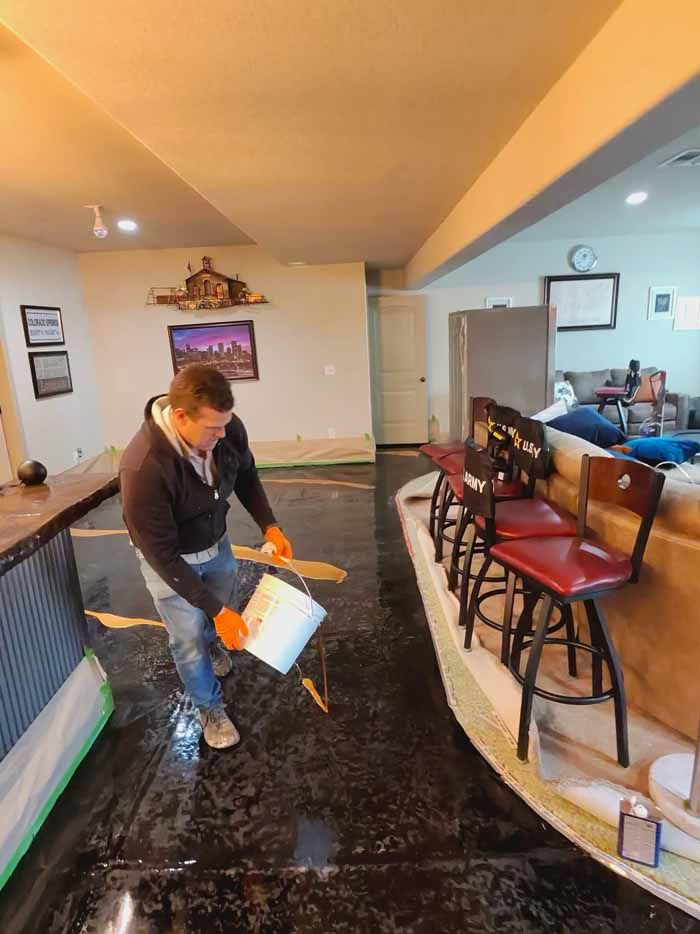 Man pouring from a bucket onto a dark floor, likely epoxy. Orange gloves, indoor setting, bar stools nearby.