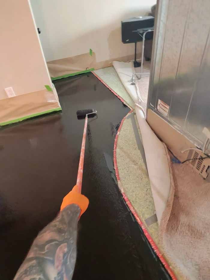 Person using paint roller on a dark floor with orange gloved hand, inside a room.