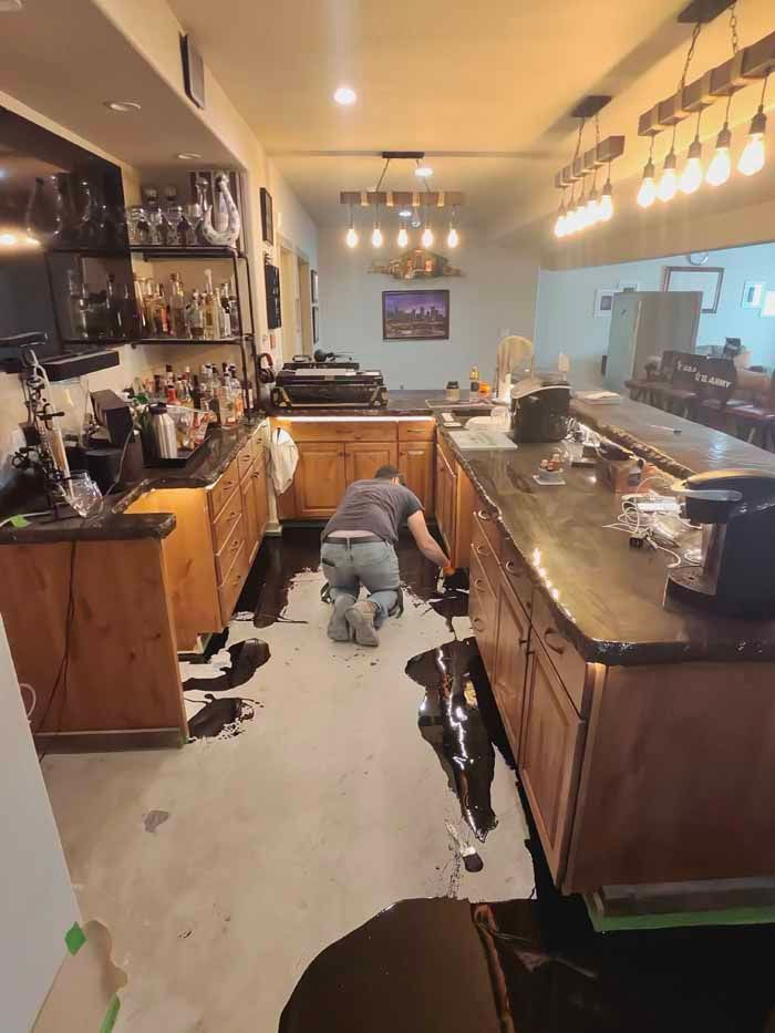Person on knees cleaning black liquid spilled on floor of a bar with dark counters, wooden cabinets, and lights overhead.