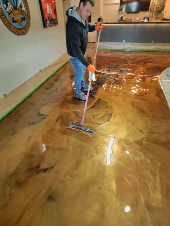 Man applying shiny, golden epoxy floor coating with a squeegee in a finished room.