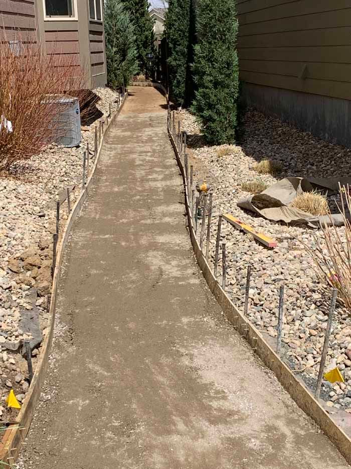 Concrete walkway under construction, bordered by wood and metal stakes. Gravel and landscaping.