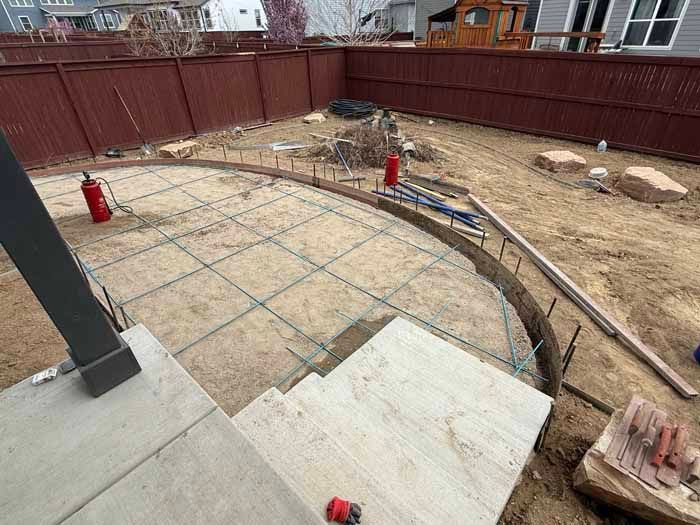 Backyard concrete slab construction: forms, rebar, and tools laid out for pouring, next to wooden fence.
