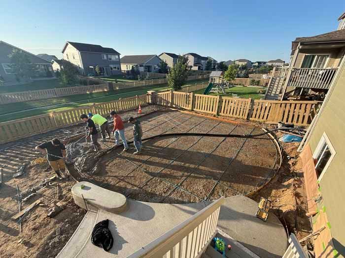 Workers pouring concrete for a circular patio in a backyard with a fence, houses, and stairs.