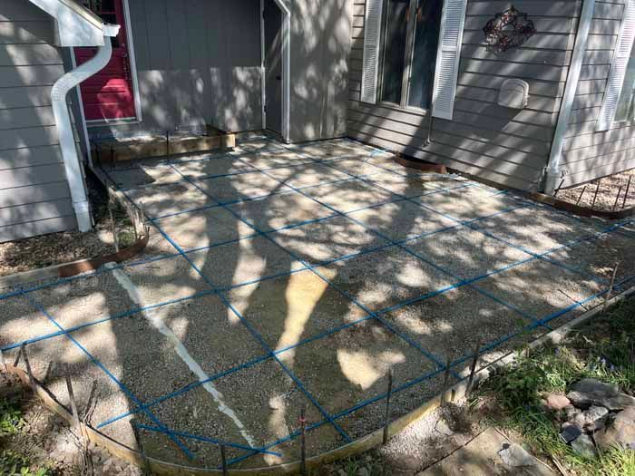 Concrete patio under construction with blue rebar grid. Grey siding and red door in background.