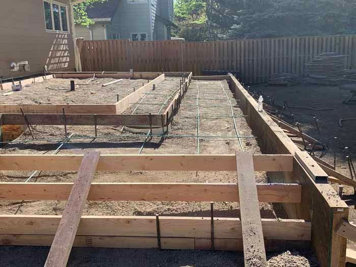 Wooden forms and rebar frame a concrete foundation in a backyard, ready for pouring.