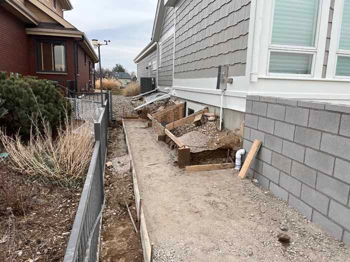 A narrow construction area beside a house, featuring gravel, a fence, and exposed foundation.