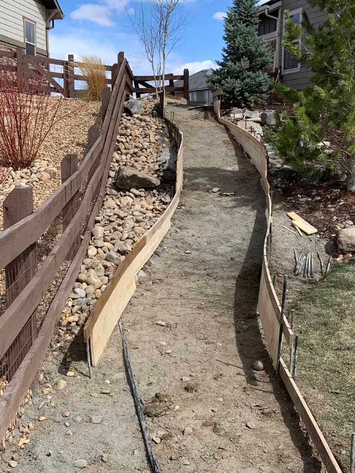 A path under construction. Wooden forms line both sides. Dirt path is between. Fences and trees are visible.