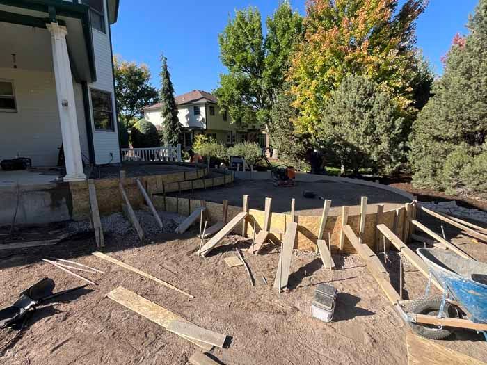 Construction site with wooden forms for a curved structure. Soil ground with a house and trees in the background.