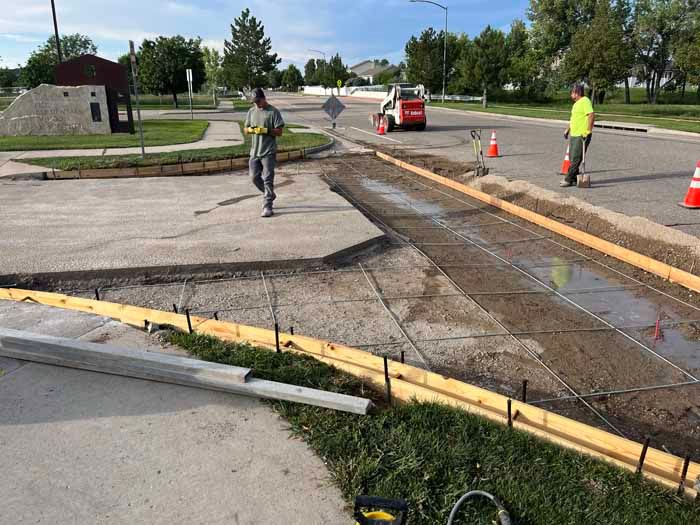 Workers pouring concrete on a road. Formwork, rebar, and orange cones visible.