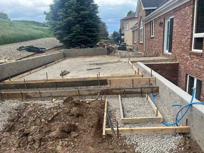 Construction site, exposed concrete foundation, wooden forms with rebar, gravel base, beside a brick building.