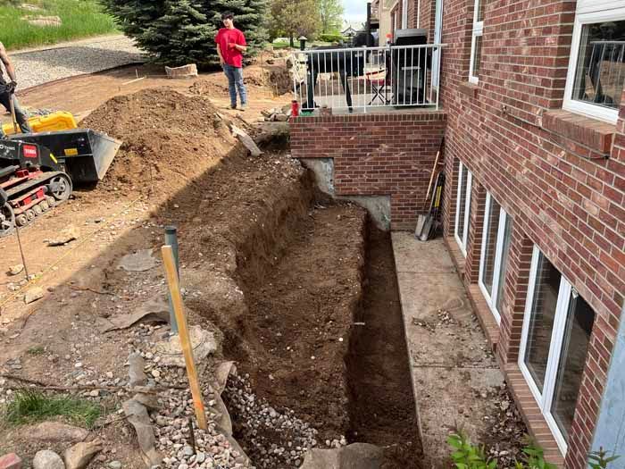 Workers excavating alongside a brick building, creating a deep trench. Soil, tools, and a small vehicle are present.