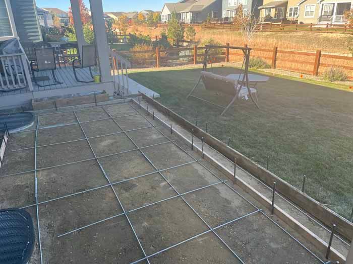 Construction site with a concrete slab frame and metal grid, set outdoors near a deck and grassy yard.