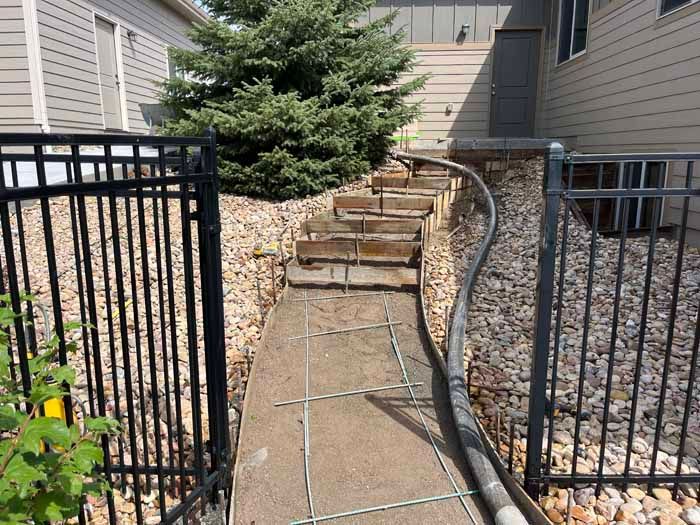 Pathway with concrete and rebar leading to steps up a small hill, bordered by fences.