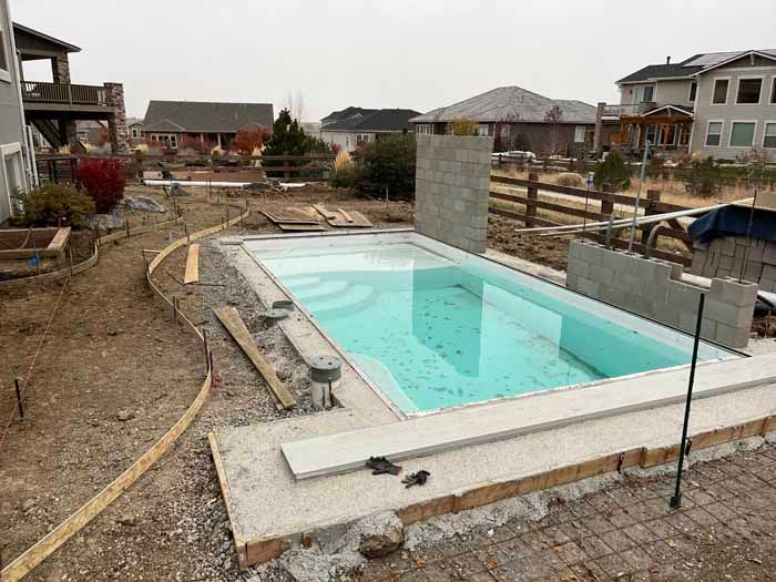 A rectangular swimming pool under construction in a residential backyard. Gray concrete and surrounding dirt are visible.