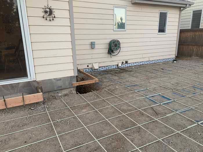Exterior view of a house with concrete foundation work. Rebar grid laid out on dirt, hole in the ground.