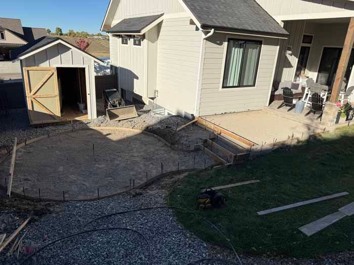 Construction site with house, shed, and curved concrete patio. Gravel and grass surround.