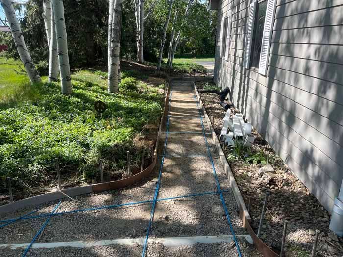 Pathway under construction with wooden forms and gravel base next to a building and trees.