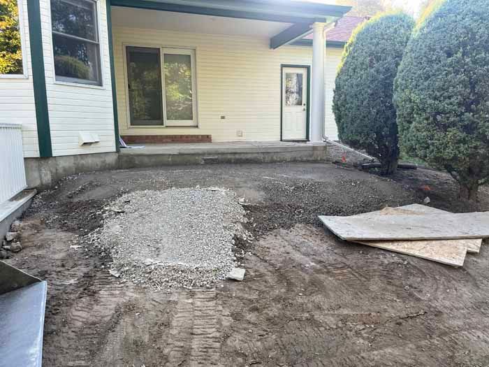 Backyard with dirt, gravel, and construction materials near a house with a porch and bushes.