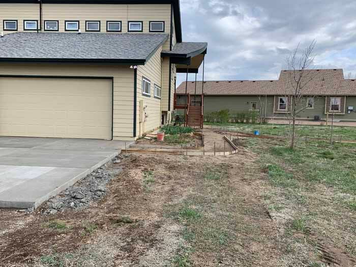 Beige house with concrete driveway, adjacent dirt area, and wooden porch steps. Another building in the background.