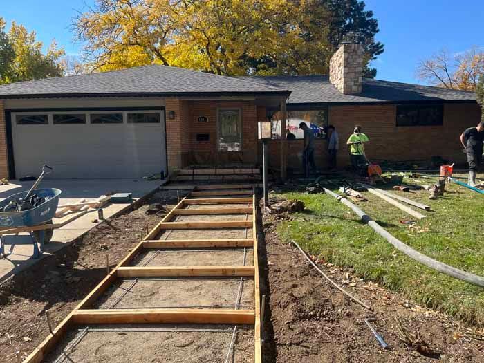 Concrete sidewalk construction in progress at a house. Workers, tools, and materials visible. Sunny day.